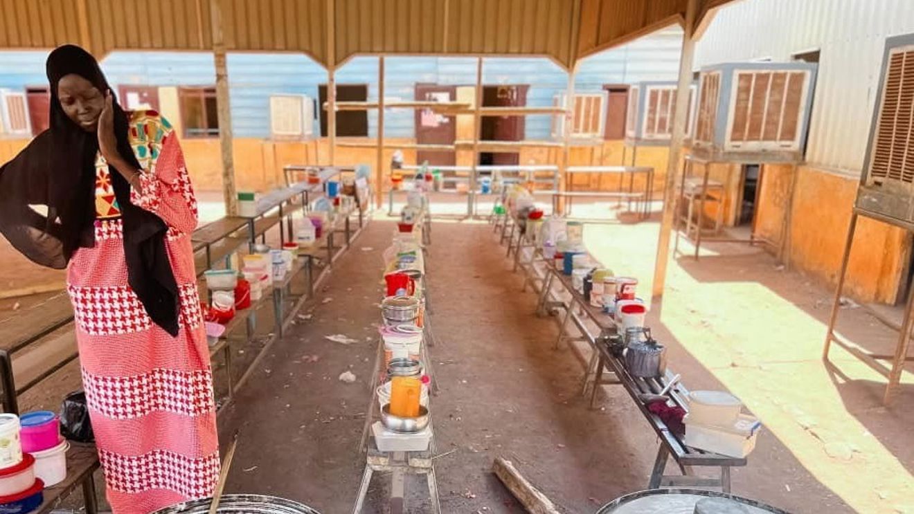 Bildet viser en kvinne i lang, rød- og hvitmønstret kjole og svart sjal som står ved en stor gryte over åpen ild. Gryten står på bakken, og det er en stor tresleiv oppi. Rundt henne er det benker og bord med mange fargerike plastbokser og kjeler, som trolig inneholder mat eller ingredienser.