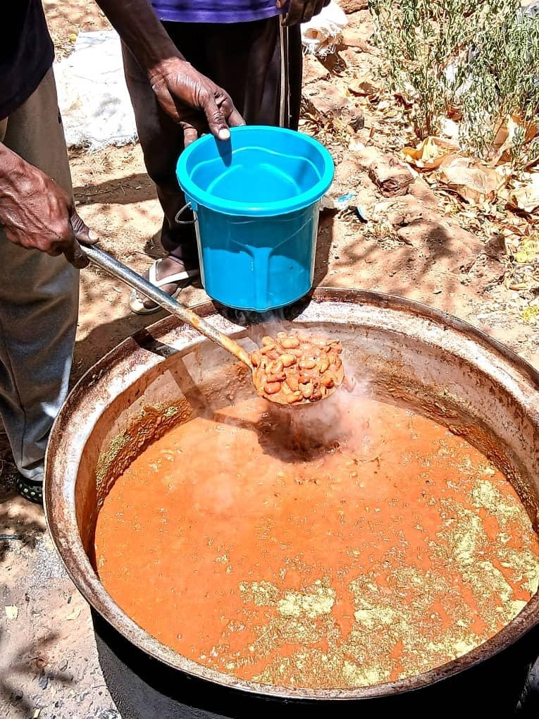 Bildet viser en stor gryte med rødbrun gryterett eller suppe som koker utendørs. En person holder en øse med bønner eller kjøtt over gryten, og en annen person holder en blå plastbøtte klar til å få mat