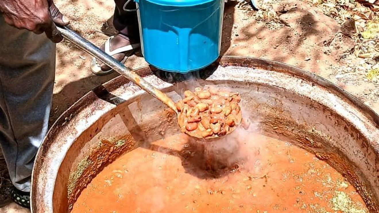 The image shows a large pot of reddish-brown stew or soup boiling outdoors. One person is holding a ladle with beans or meat over the pot, and another person is holding a blue plastic bucket ready to receive food.