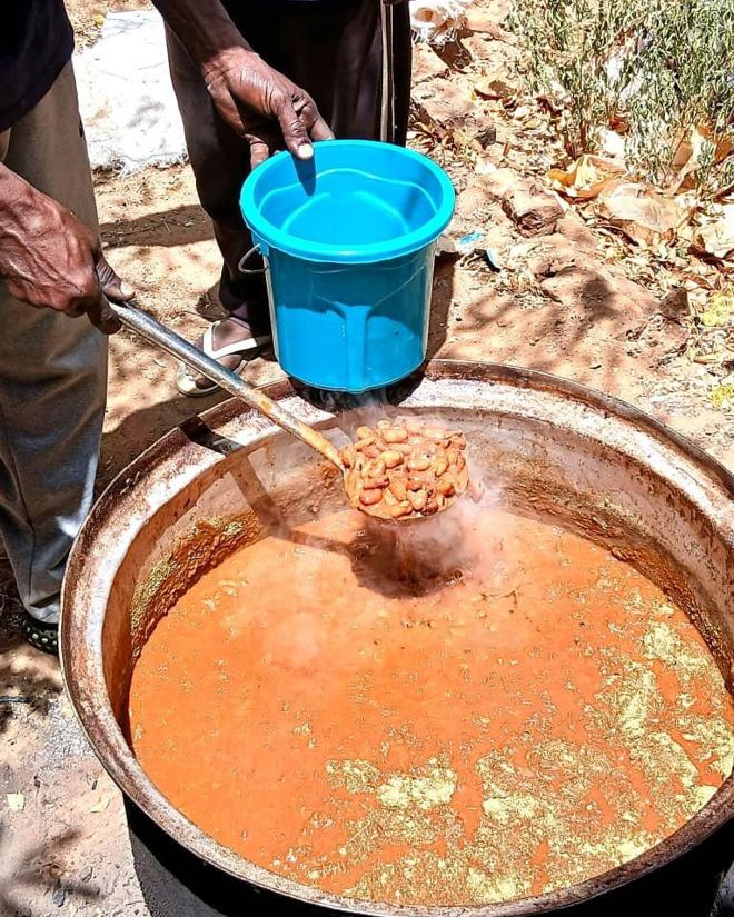 Bildet viser en stor gryte med rødbrun gryterett eller suppe som koker utendørs. En person holder en øse med bønner eller kjøtt over gryten, og en annen person holder en blå plastbøtte klar til å få mat