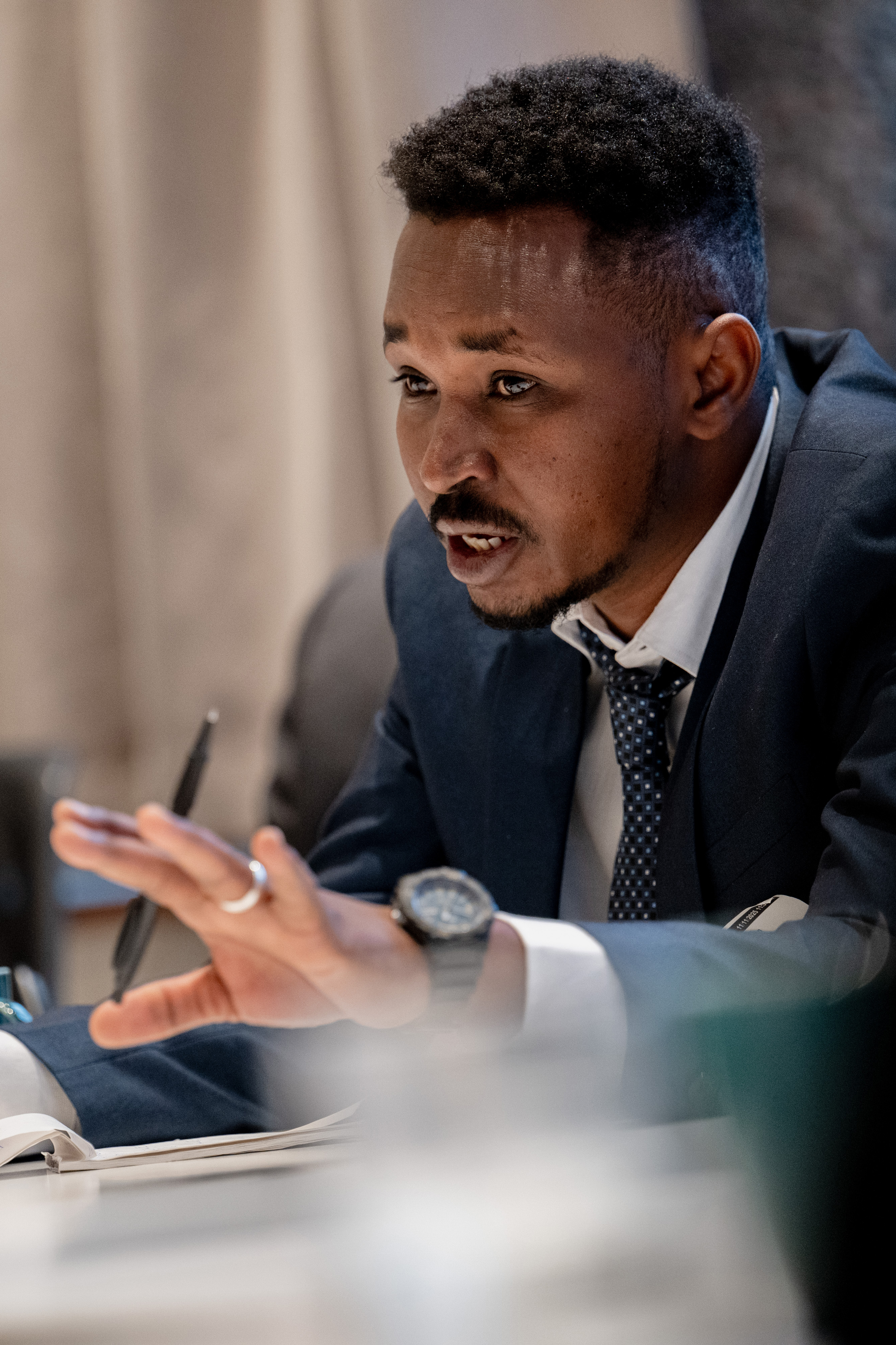 En ung mann fra Sudan sitter ved et bord iført dress og slips. Han holder en penn og har hånden løftet, som om han deltar aktivt i en samtale eller et møte. På bordet foran ham ligger et åpent notatark.