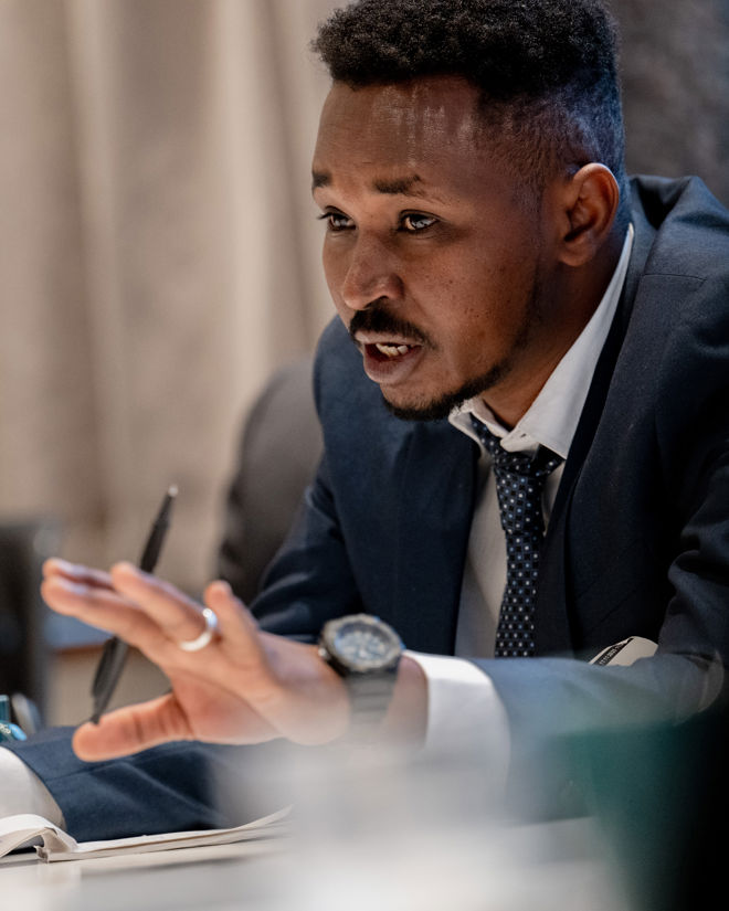 En ung mann fra Sudan sitter ved et bord iført dress og slips. Han holder en penn og har hånden løftet, som om han deltar aktivt i en samtale eller et møte. På bordet foran ham ligger et åpent notatark.