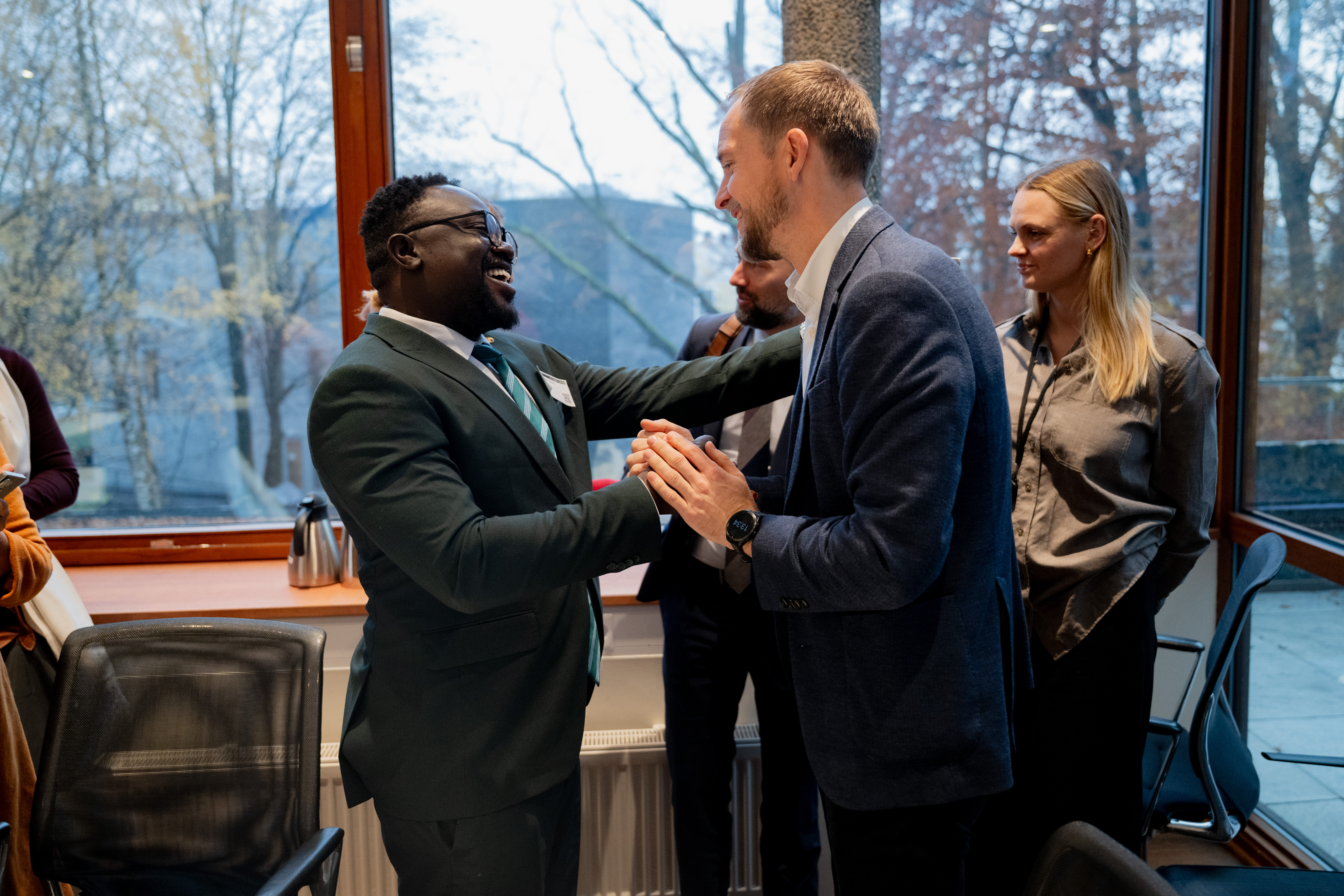  To menn står nær hverandre midt i rommet og hilser eller småprater med et håndtrykk og kroppskontakt, begge kledd i dress.