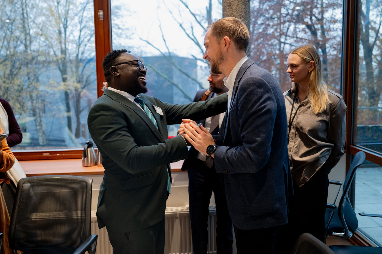  To menn står nær hverandre midt i rommet og hilser eller småprater med et håndtrykk og kroppskontakt, begge kledd i dress.