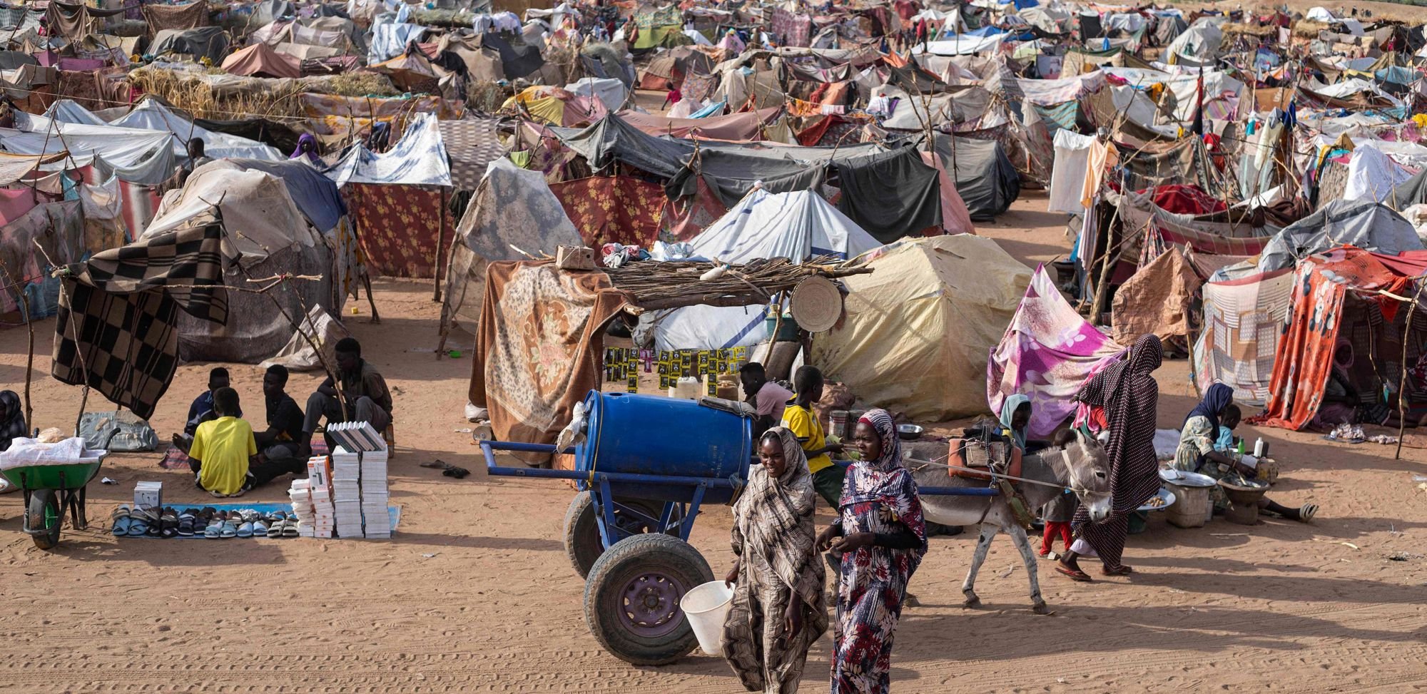 Bildet viser en stor flyktningleir i Sudan. I forgrunnen går tre kvinner i fargerike kjoler på sandgrunn. Rundt dem står mange provisoriske telt laget av tepper og presenninger i ulike farger og mønstre. Flere voksne og barn sitter eller beveger seg mellom teltene. Det er et esel med kjerre, og noen selger varer på bakken. I bakgrunnen sees ørkenlandskap, spredte trær og fjell.
