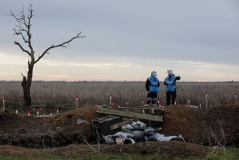 Fotografi av et landskap med en enslig, bladløs tre til venstre og to personer i blå beskyttelsesvester til høyre. De står ved et område markert med rødt og hvitt, sannsynligvis et mineryddingsområde. Bakgrunnen viser en vidstrakt eng under en overskyet himmel.