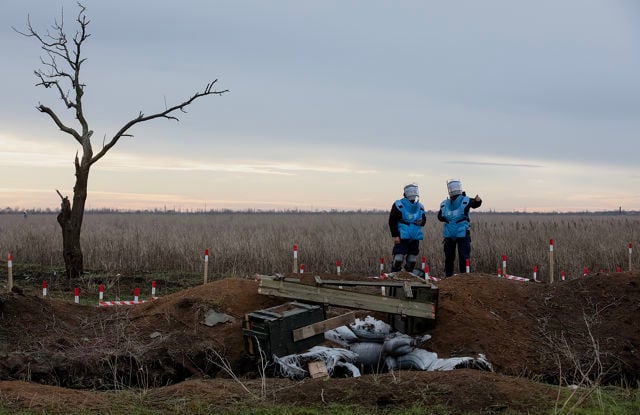 The image shows two individuals wearing protective blue vests and helmets, standing in a field marked with red-and-white sticks, likely indicating a restricted or hazardous area. The foreground includes a small wooden bridge over a trench filled with sandbags.