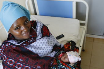 En kvinne sitter på en sykehusseng med et spedbarn svøpt i et fargerikt klede i armene.