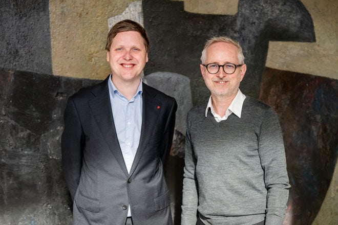 The image features two individuals standing side by side smiling in front of a textured wall that displays abstract art consisting of dark and light shades.