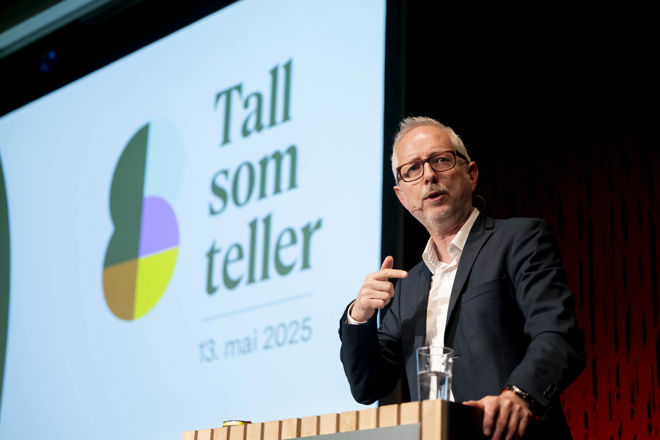 The image shows a person standing at a podium, seemingly giving a presentation or speech. The background features a large screen displaying the text "Tall som teller" along with the date "13. mai 2025." 