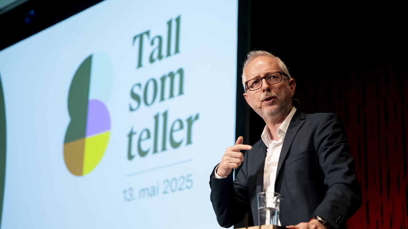 Bildet viser direktø i Norad Bård Vegard Solhjell som står ved en talerstol foran en stor skjerm som viser teksten "Tall som teller" med datoen "13. mai 2025".