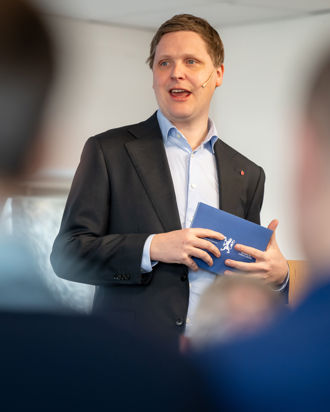 En person i dressjakke og lyseblå skjorte står og holder et blått hefte med det norske riksvåpenet på forsiden. Personen ser ut til å være i en formell setting, for eksempel et møte eller en presentasjon, og det er andre mennesker til stede i rommet, synlige som uklare silhuetter i forgrunnen. Stemningen er profesjonell og seriøs.