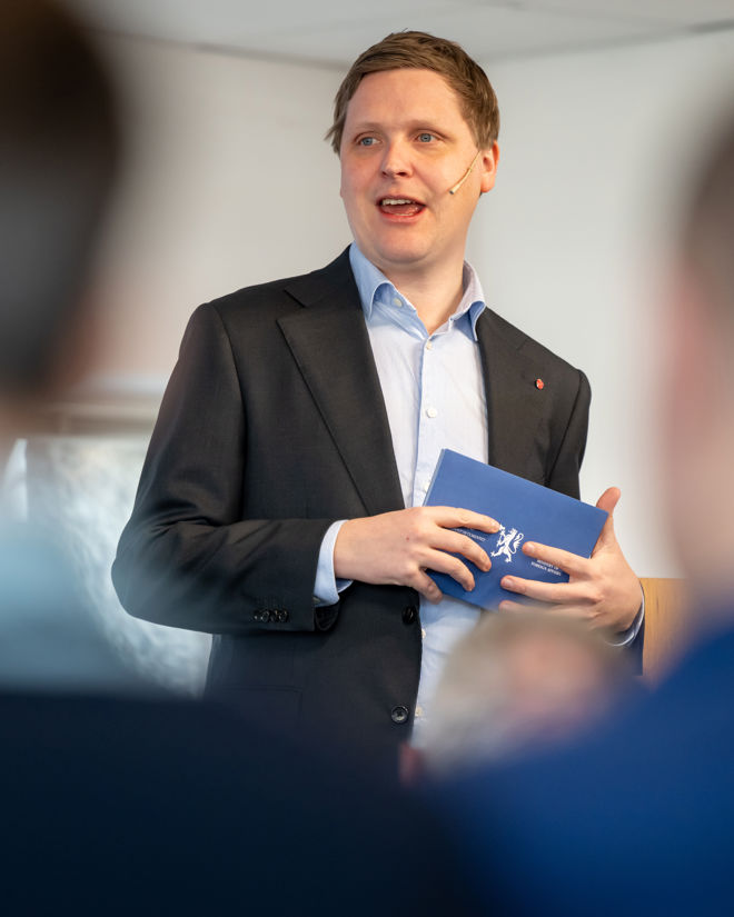 En person i dressjakke og lyseblå skjorte står og holder et blått hefte med det norske riksvåpenet på forsiden. Personen ser ut til å være i en formell setting, for eksempel et møte eller en presentasjon, og det er andre mennesker til stede i rommet, synlige som uklare silhuetter i forgrunnen. Stemningen er profesjonell og seriøs.