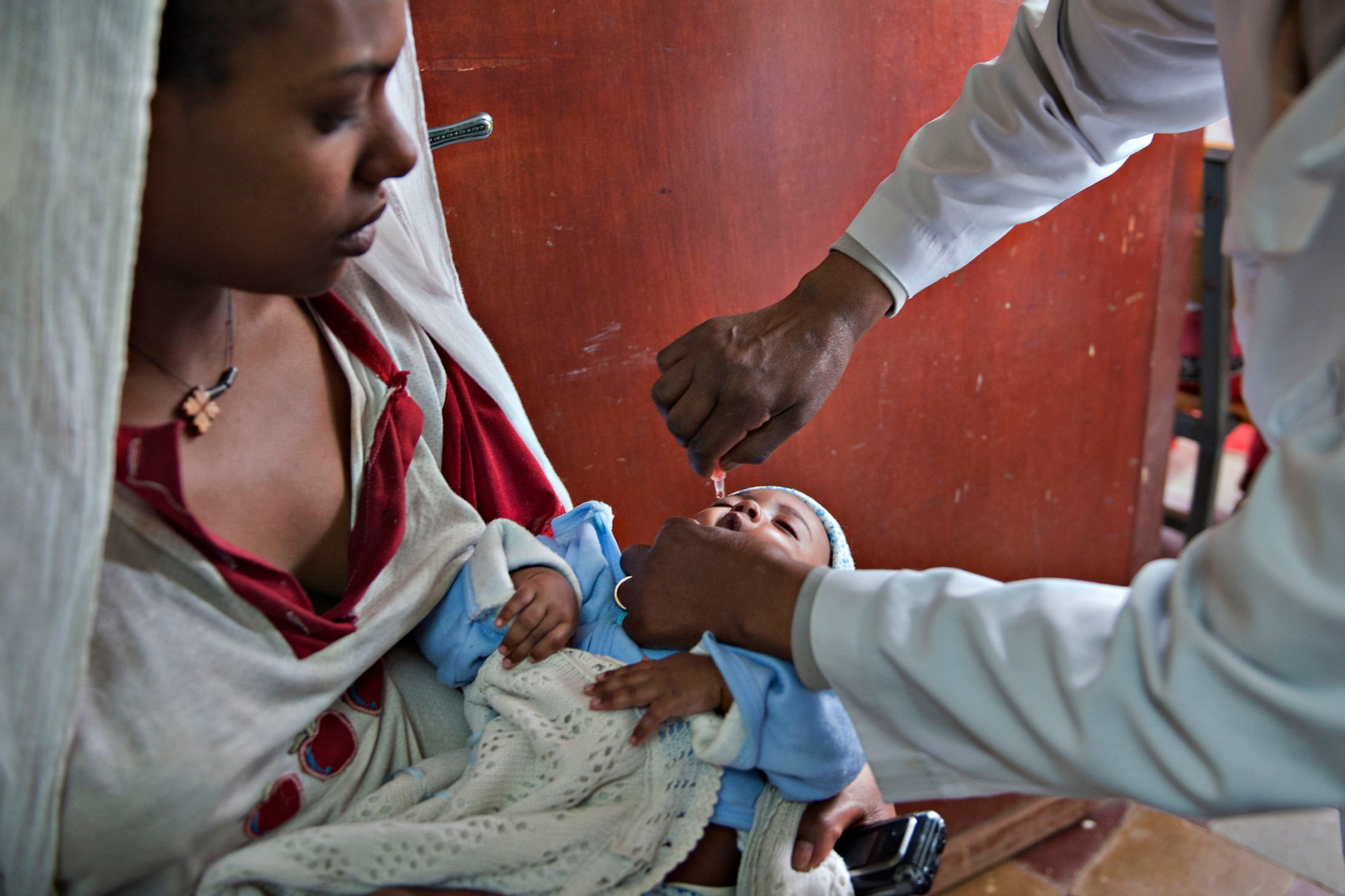 The image shows an adult holding a small child in their arms. The child is wrapped in a light-colored blanket and dressed in blue. Another person, wearing a white coat, administers a drop of liquid from a pipette—likely a vaccine or medical treatment. The scene takes place indoors, in front of a brown wall or door. The atmosphere suggests a healthcare setting where the child’s well-being is being cared for.