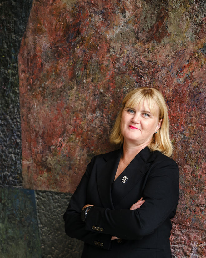 A woman with shoulder-length blonde hair stands with her arms crossed in front of a large, textured and colorful wall. She is dressed in a dark blazer with a decorative pin on the lapel. The atmosphere is formal and confident.