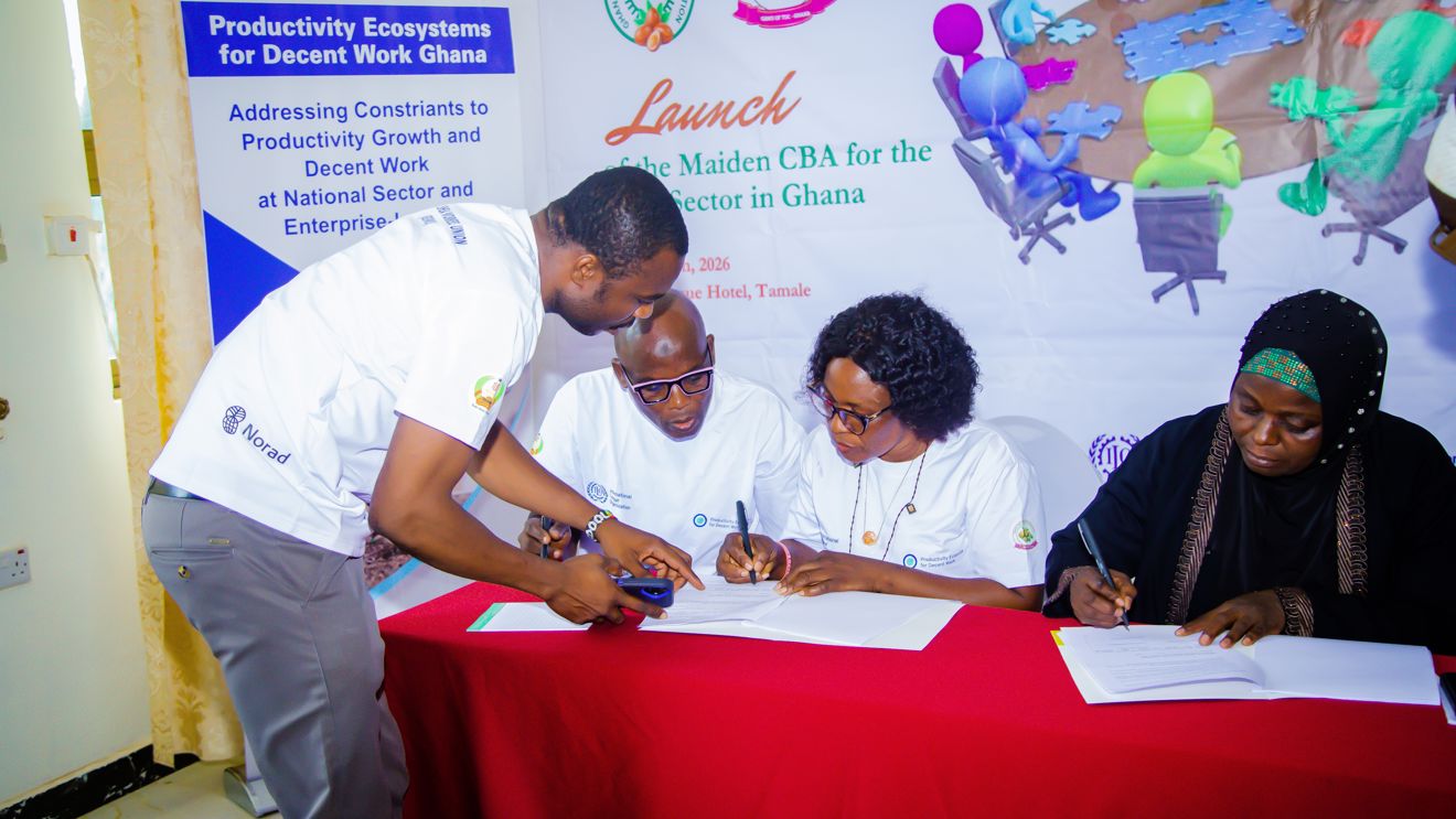 Four adults sit at a table signing documents during an event, with banners about productivity and decent work in the background.