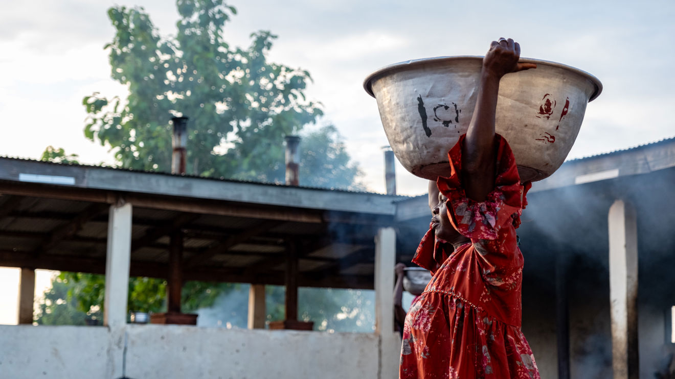 A woman in a red dress carries a large bowl on her head. She is outdoors and the sky is blue.
