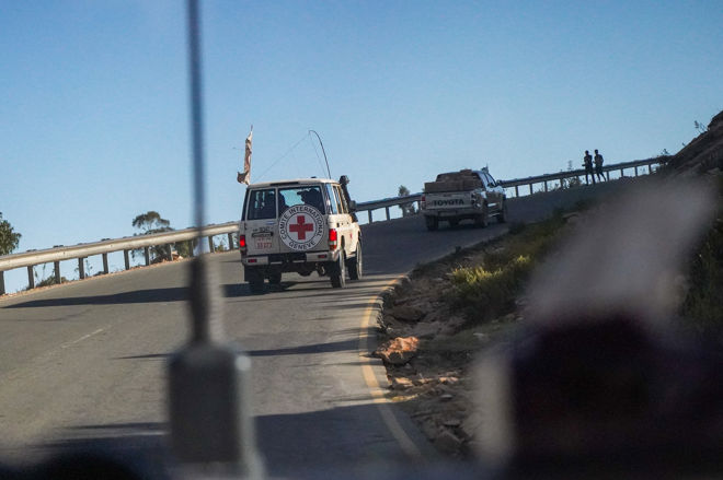 Bildet viser et kjøretøy fra Røde Kors og en pickup på en svingete vei, med noen personer stående ved autovernet i bakgrunnen.