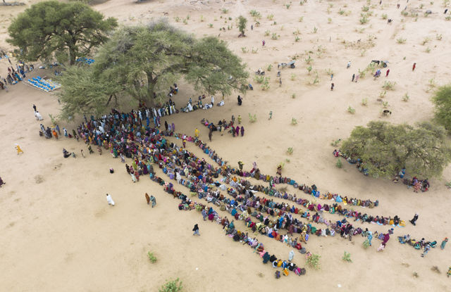 Dette bildet viser en stor gruppe mennesker samlet i et åpent, tørt område. De fleste av dem sitter i lange, parallelle rekker som strekker seg ut på bakken. Mange av dem har fargerike klær som skaper et levende mønster mot den lyse sanden.