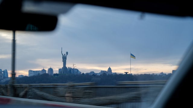 Bilde tatt fra innsiden av en bil viser bysilhuetten i Kyiv med Moderlandet-statuen og et ukrainsk flagg mot himmelen.