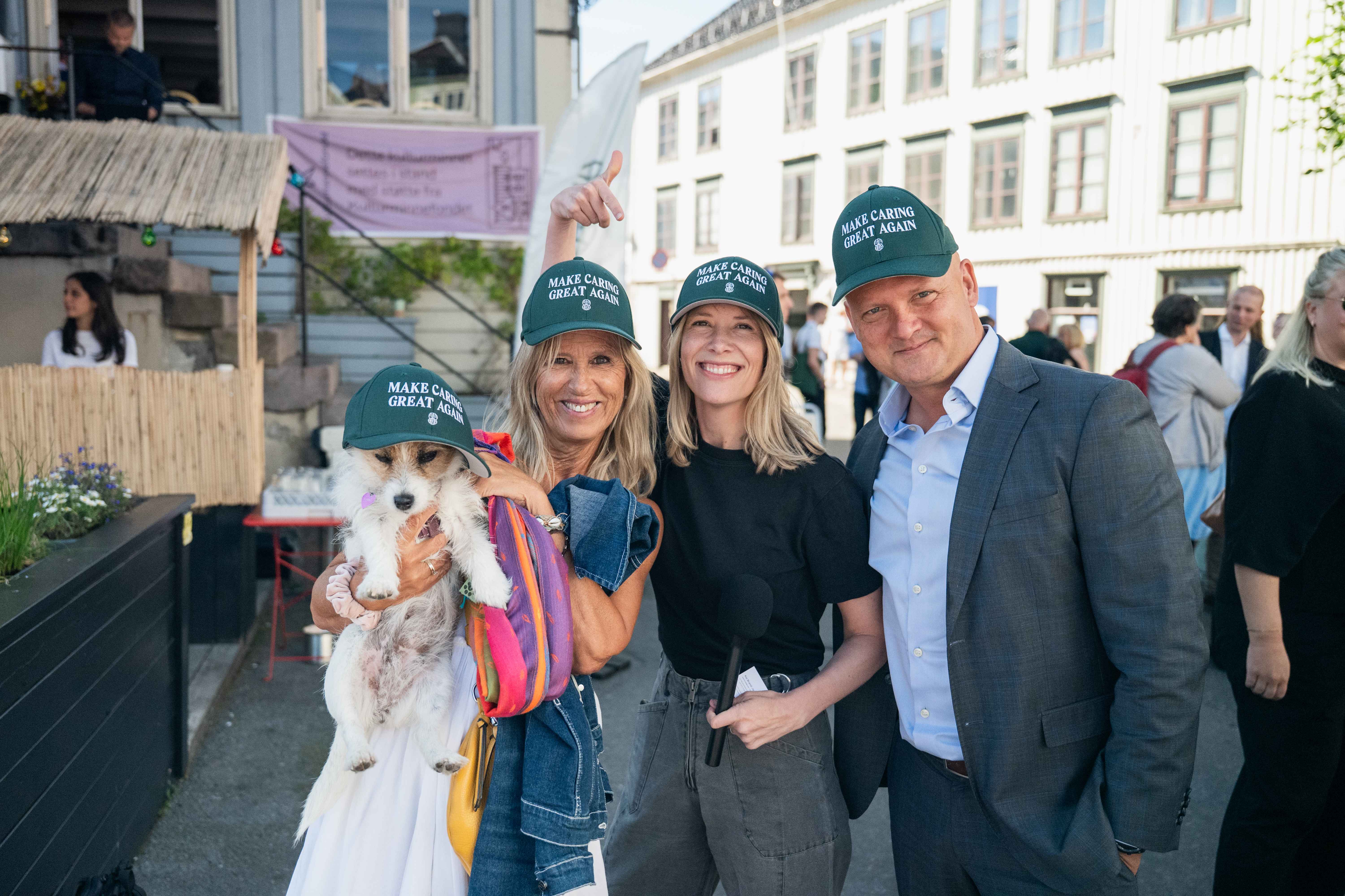 Tre voksne personer med caps står tett sammen og smiler til kamera i en gate med flere mennesker i bakgrunnen. Personen til venstre holder en liten hund med caps.