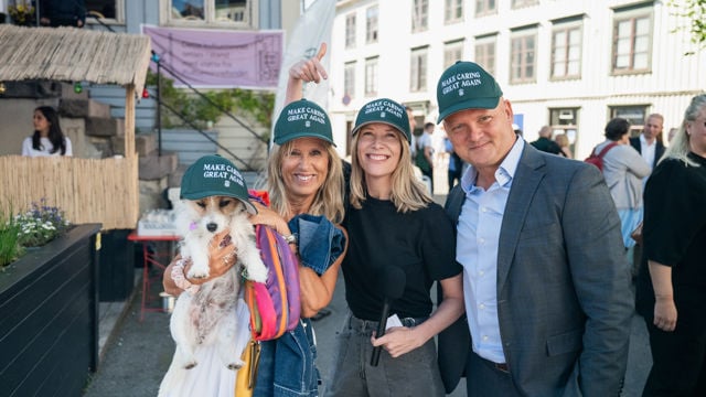 Tre voksne personer med caps står tett sammen og smiler til kamera i en gate med flere mennesker i bakgrunnen. Personen til venstre holder en liten hund med caps.