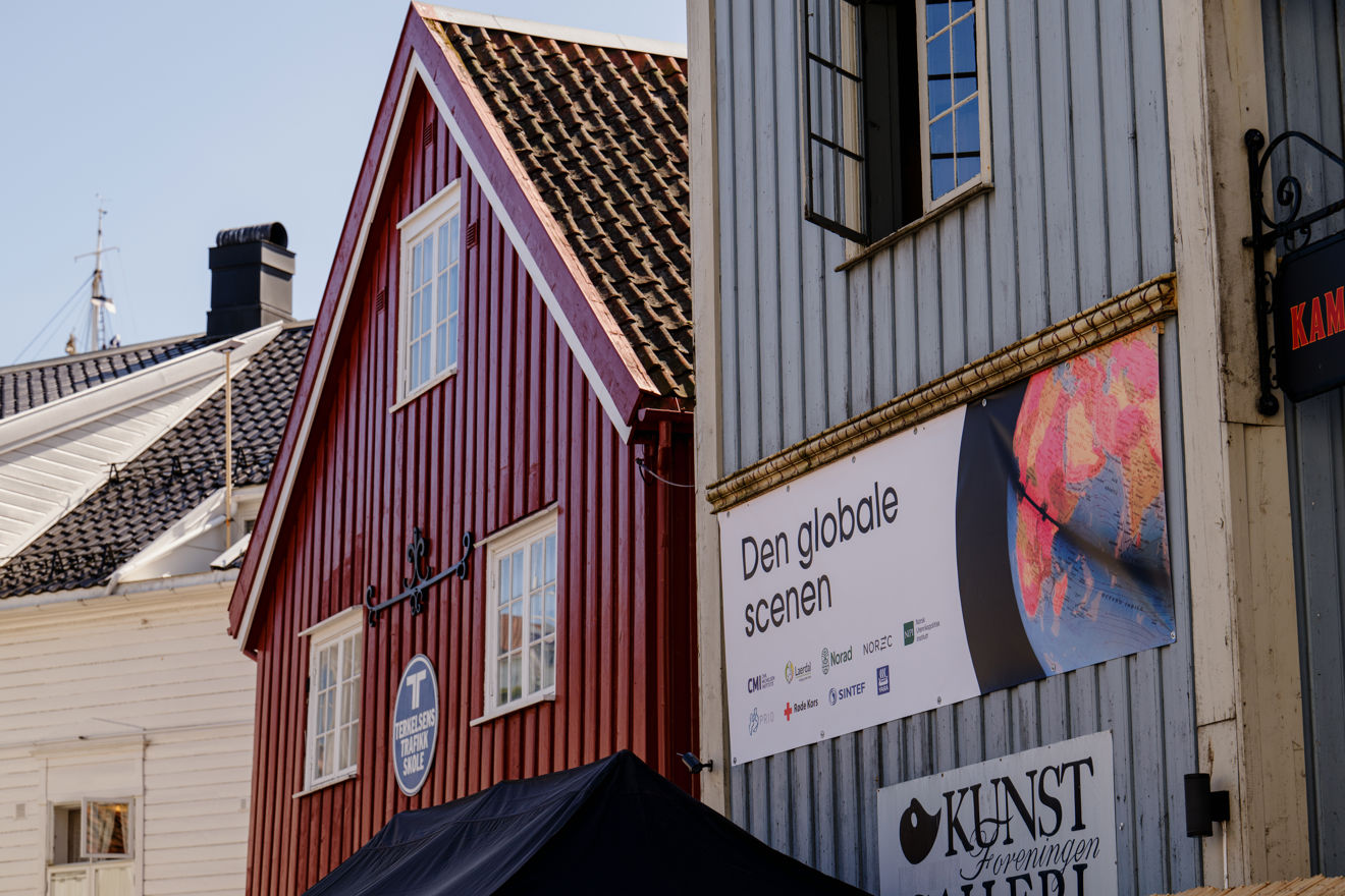 En rød bygning med hvite vinduskarmer står mellom andre hus i en gate. Til høyre i bildet henger et skilt og et rundt, fargerikt merke på veggen.