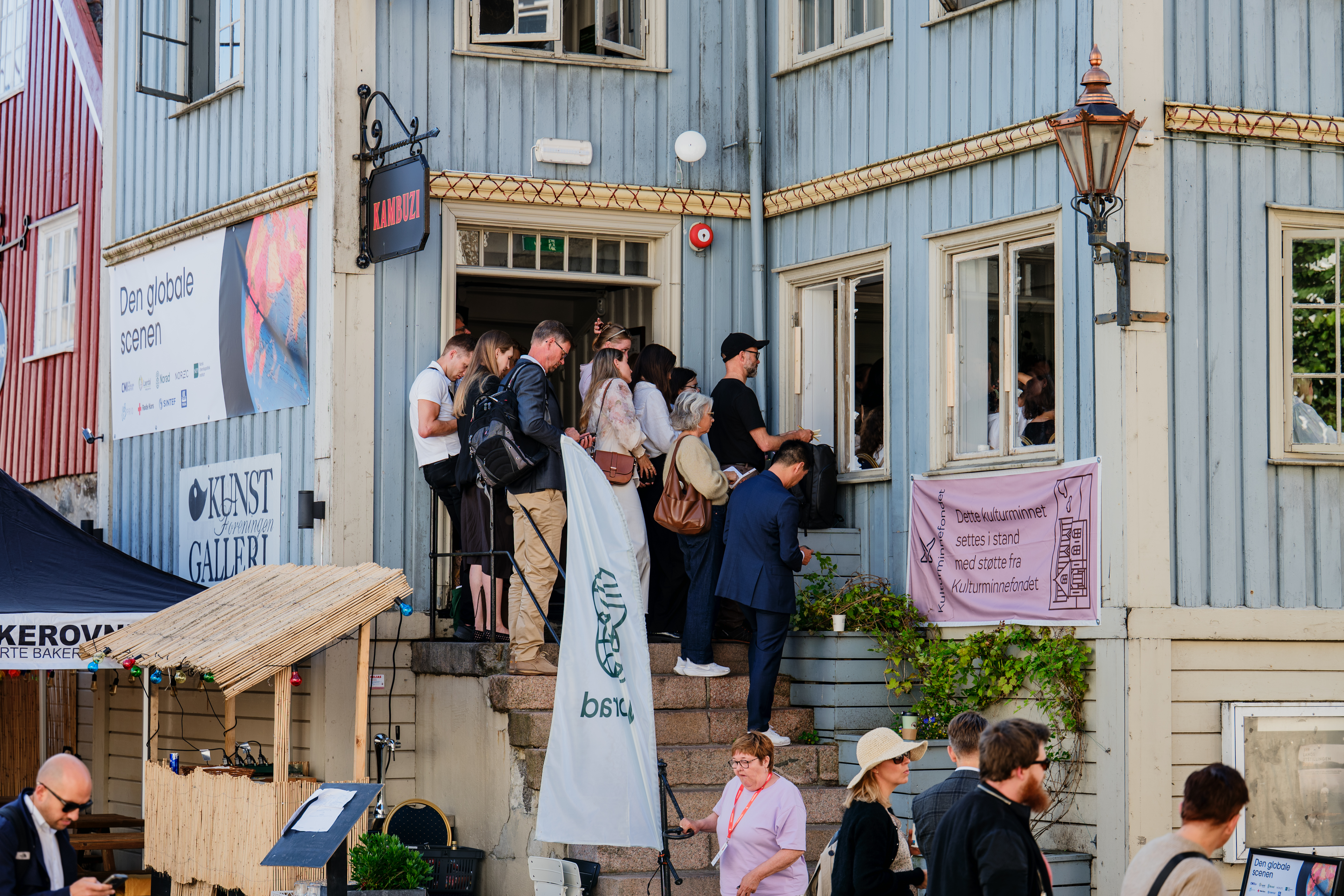 Flere voksne personer står på en trapp utenfor en bygning og ser ut mot folk som står nedenfor. Det henger et rosa banner på rekkverket, og flere personer er samlet i gaten foran bygningen.