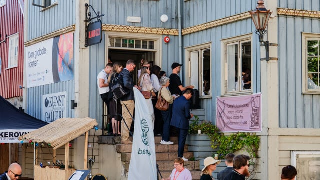 Flere voksne personer står på en trapp utenfor en bygning og ser ut mot folk som står nedenfor. Det henger et rosa banner på rekkverket, og flere personer er samlet i gaten foran bygningen.