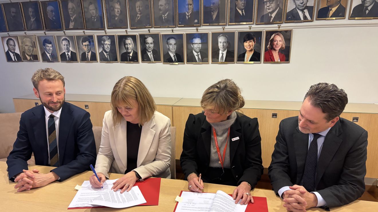 To kvinner sitter ved et bord og signerer dokumenter, med to menn i dress sittende på hver side.