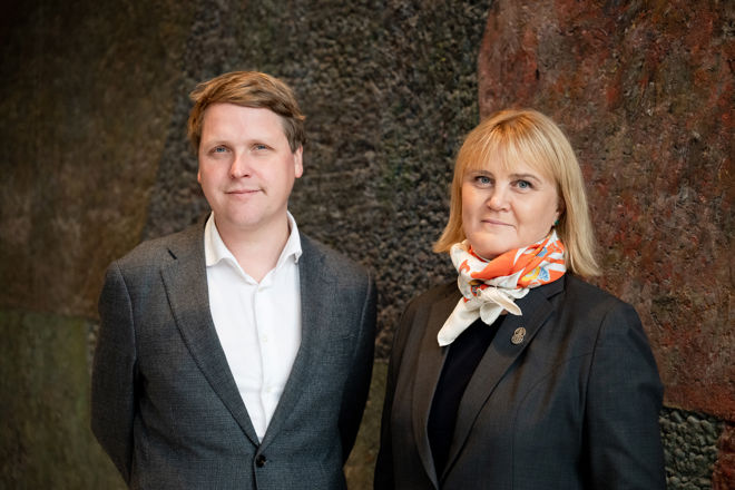 Bildet viser en mann og en kvinne som står ved siden av hverandre foran en mørk vegg. Begge er kledd i formelle klær.