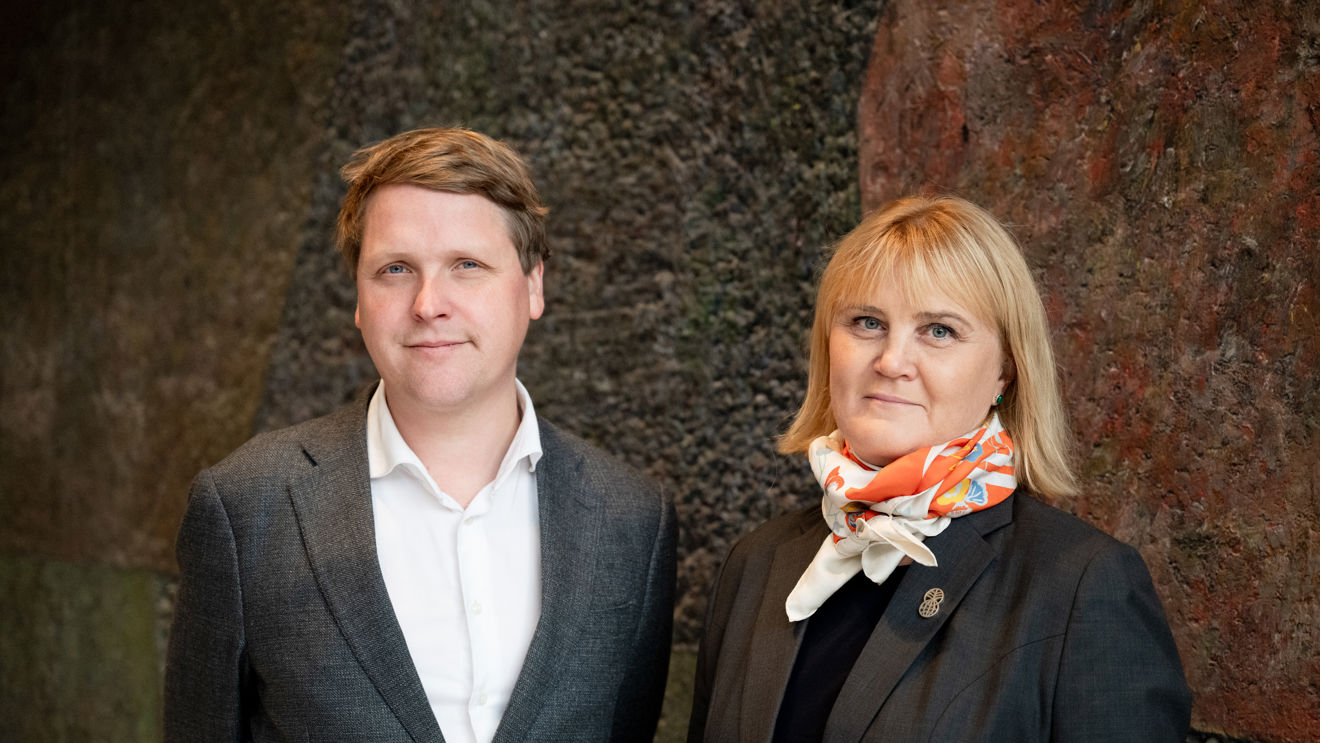 Bildet viser en mann og en kvinne som står ved siden av hverandre foran en mørk vegg. Begge er kledd i formelle klær.