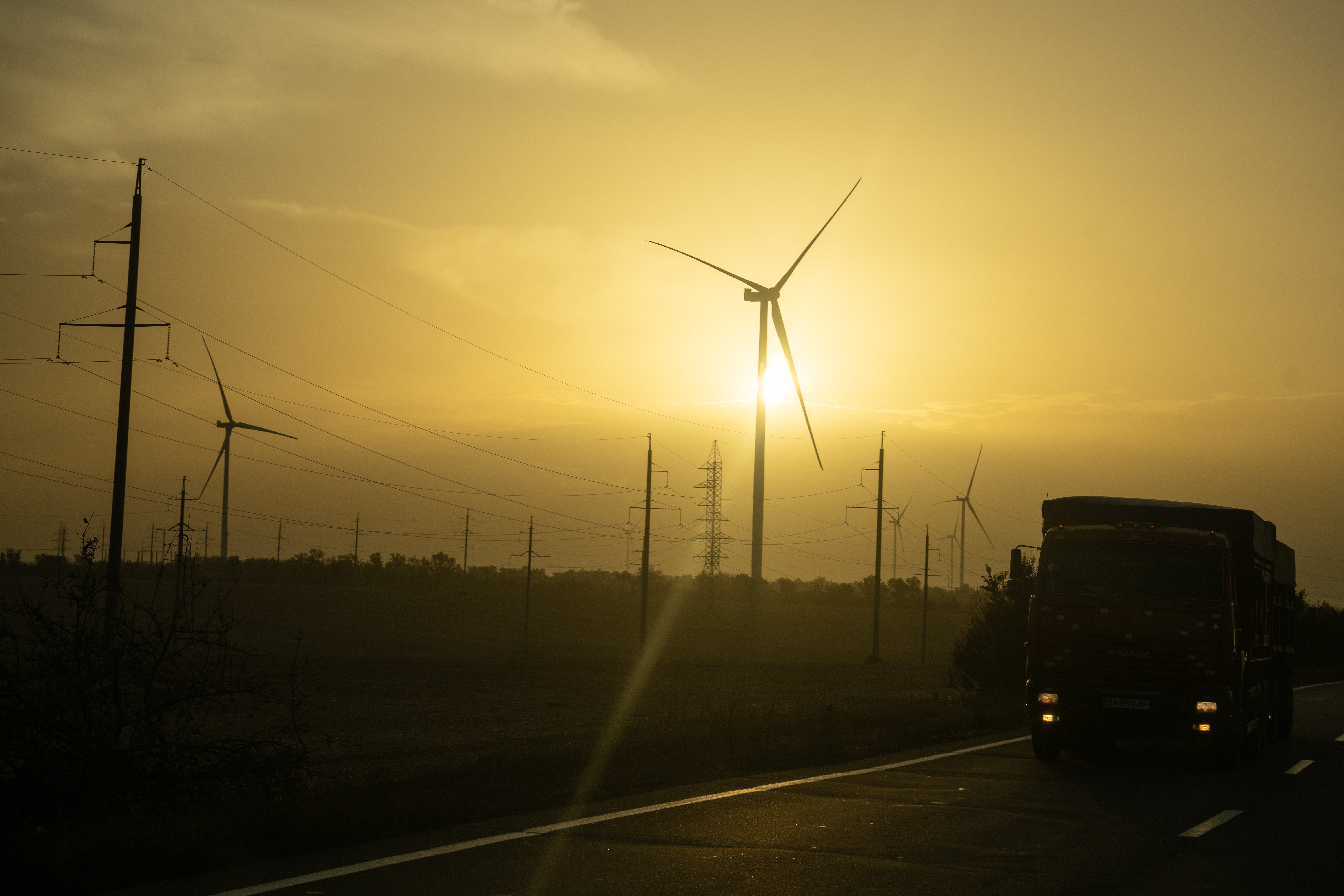 En lastebil kjører på en vei ved siden av flere vindturbiner og kraftledninger i solnedgang.