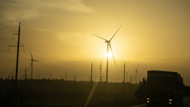 En lastebil kjører på en vei ved siden av flere vindturbiner og kraftledninger i solnedgang.