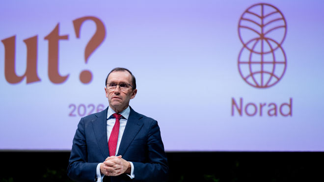 En mann i dress og rød slips står foran en skjerm med Norad-logoen og tekst i bakgrunnen.