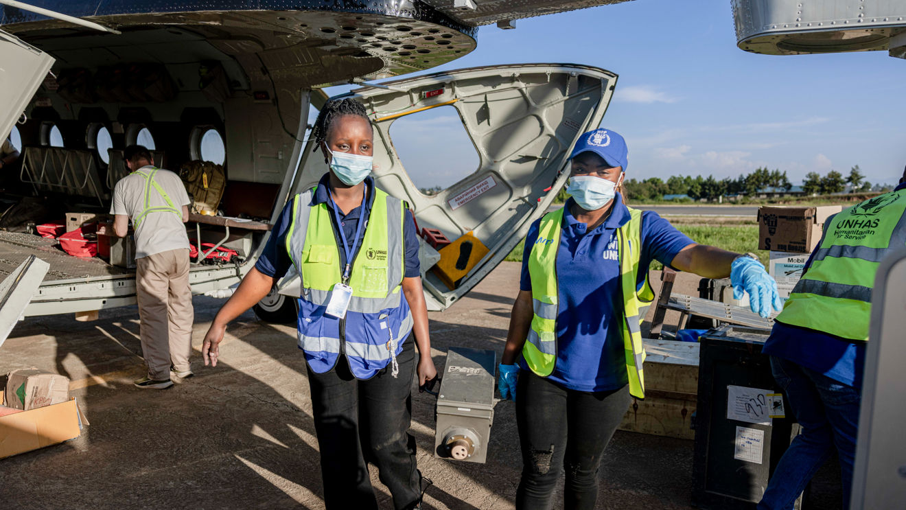 Personer i refleksvester laster av utstyr fra et fly på en flyplass. To personer bærer en kasse bort fra flyet, mens andre håndterer utstyr i bakgrunnen.