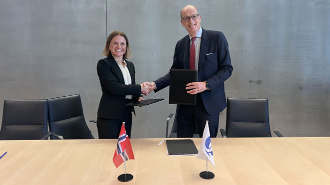 En kvinne og en mann står ved et bord og håndhilser mens de holder dokumentmapper. Norske og internasjonale flagg står på bordet foran dem.