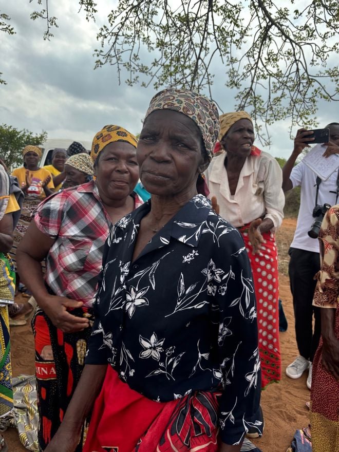 En gruppe voksne kvinner står utendørs på bar jord, flere kledd i mønstrede klær og hodeplagg, med trær og overskyet himmel i bakgrunnen.