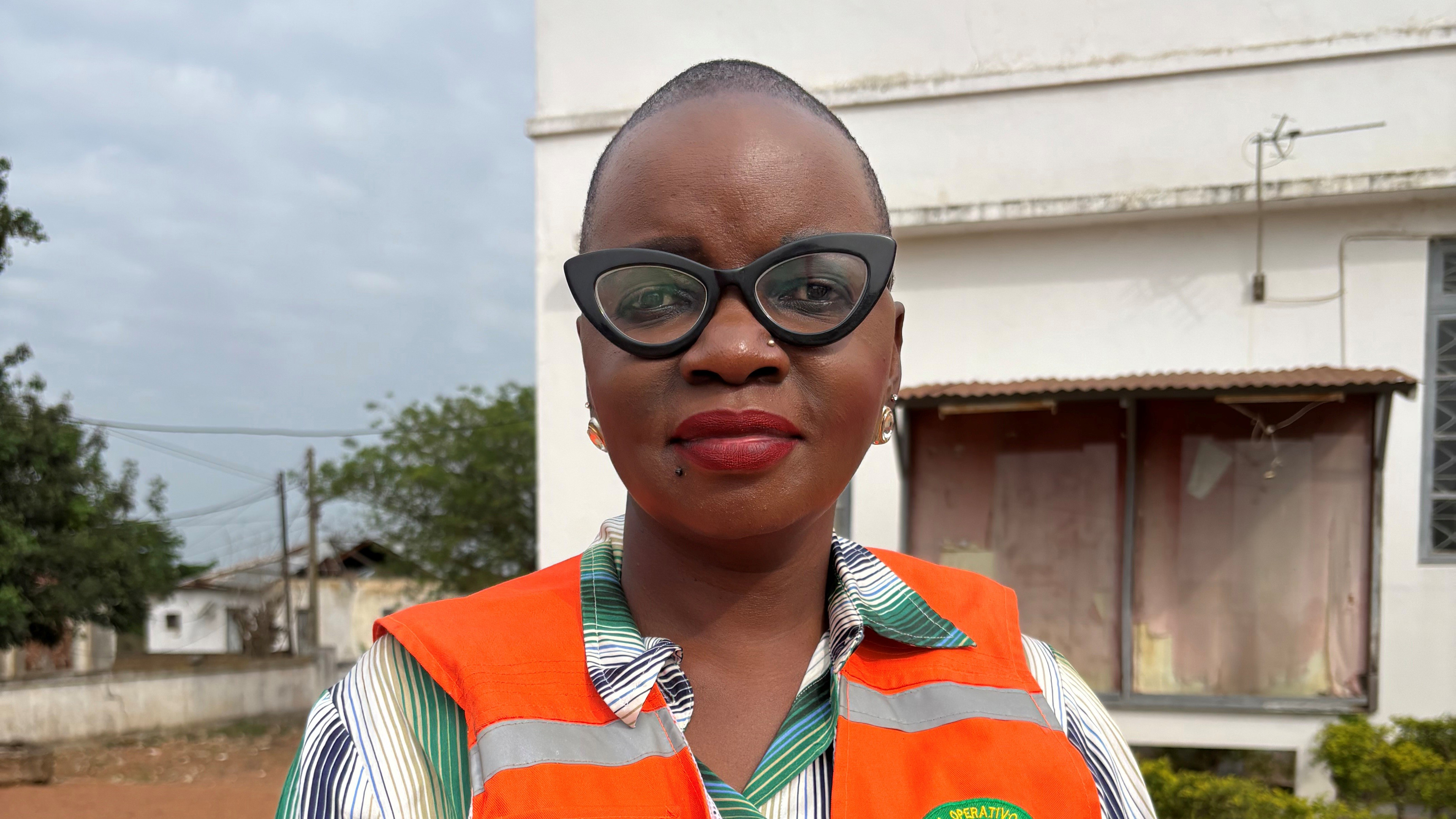 En person iført oransje refleksvest og stripete skjorte står utendørs foran en lys bygning.