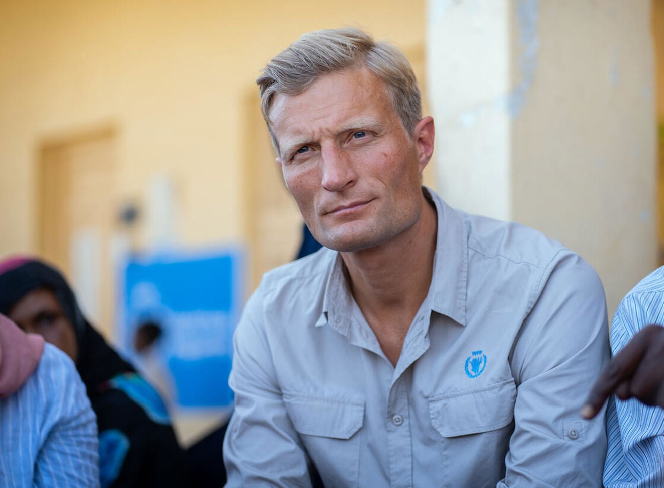 En mann i lys blå skjorte sitter innendørs og ser rolig til siden, med en uskarp bakgrunn som tyder på et møte eller en samtale.