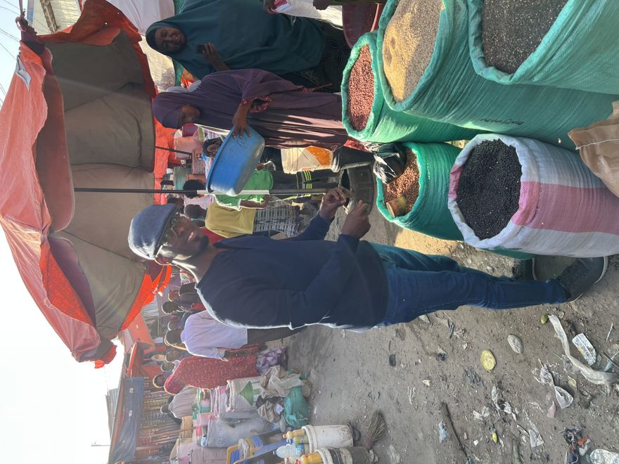  denne En mann kjøper bønner og korn fra store sekker på et utendørs marked, mens en kvinne med bøtte står bak et bord under en parasoll.