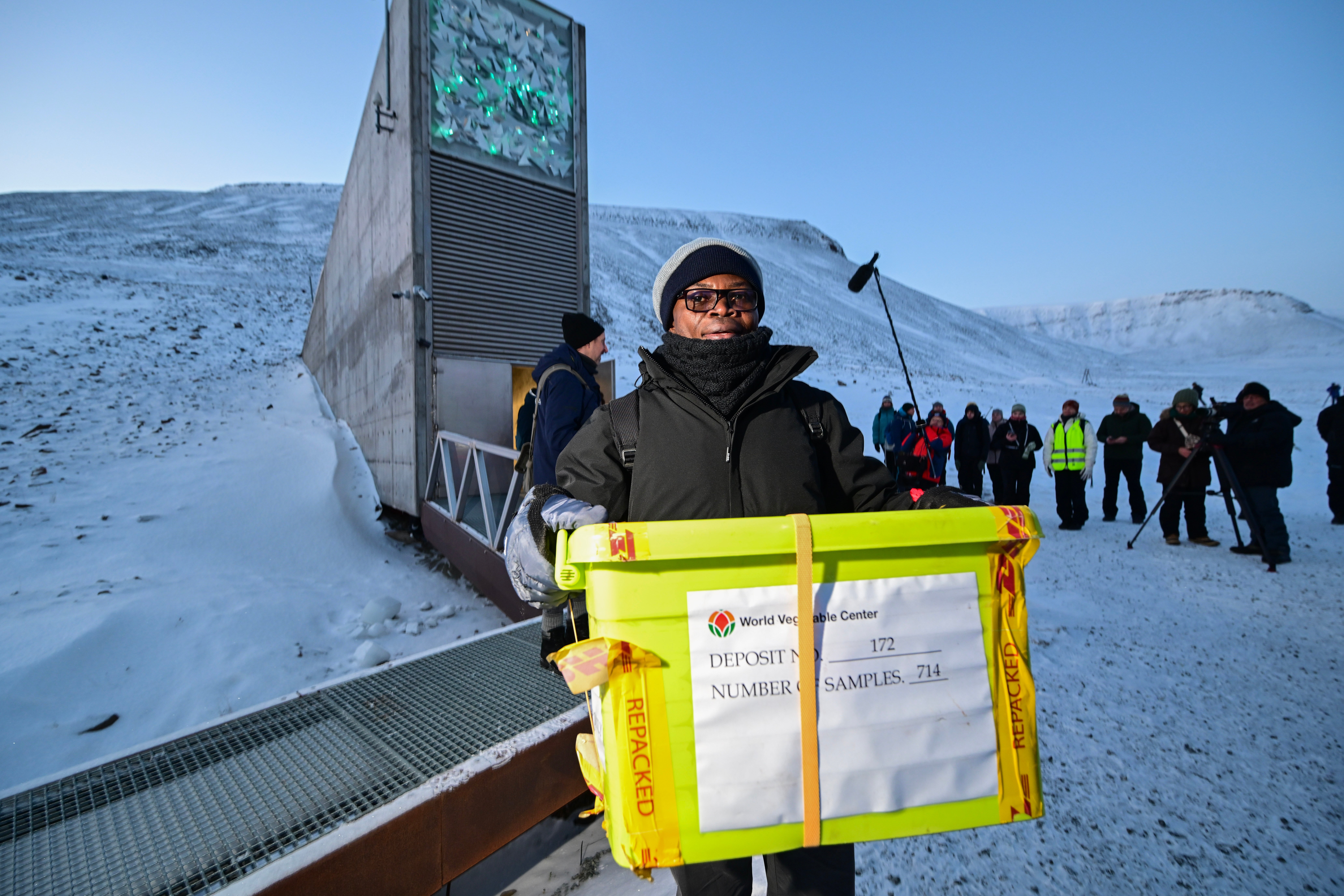 En person med vinterklær bærer en stor plastkasse foran inngangen til Svalbard globale frøhvelv, mens flere mennesker står og ser på i bakgrunnen i snødekt landskap.