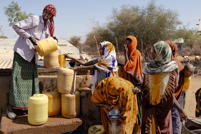 En gruppe kvinner og barn fyller gule plastkanner med vann fra en brønn i et tørt utendørsmiljø.