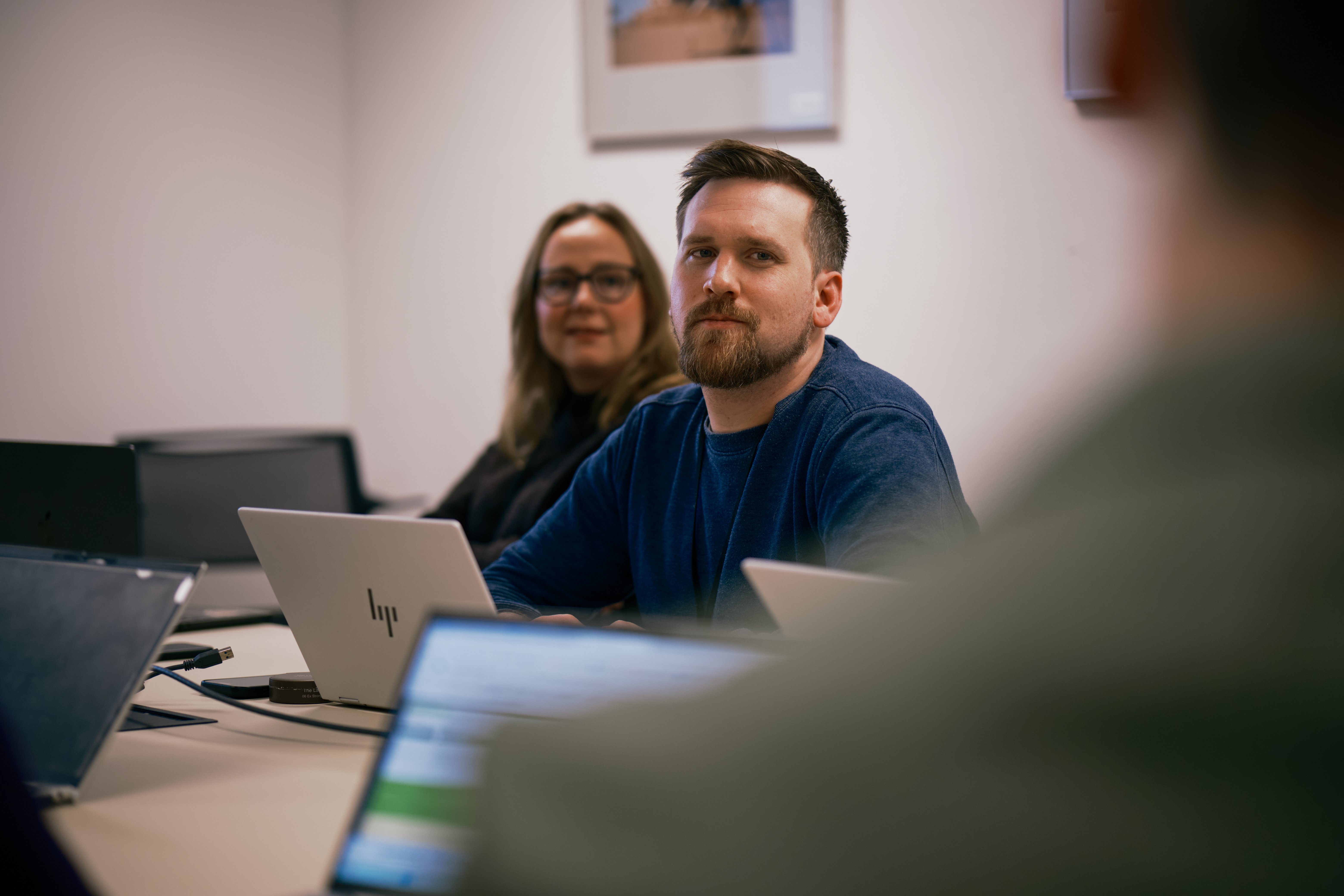 Two young adults sit at a meeting table with open laptops, while another person is partially visible in the foreground; a framed picture hangs on the wall behind them.