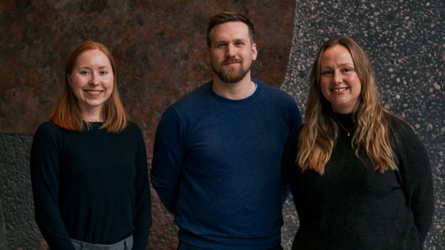 Three young adults stand side by side in front of an abstract patterned wall, all wearing dark clothing and smiling.