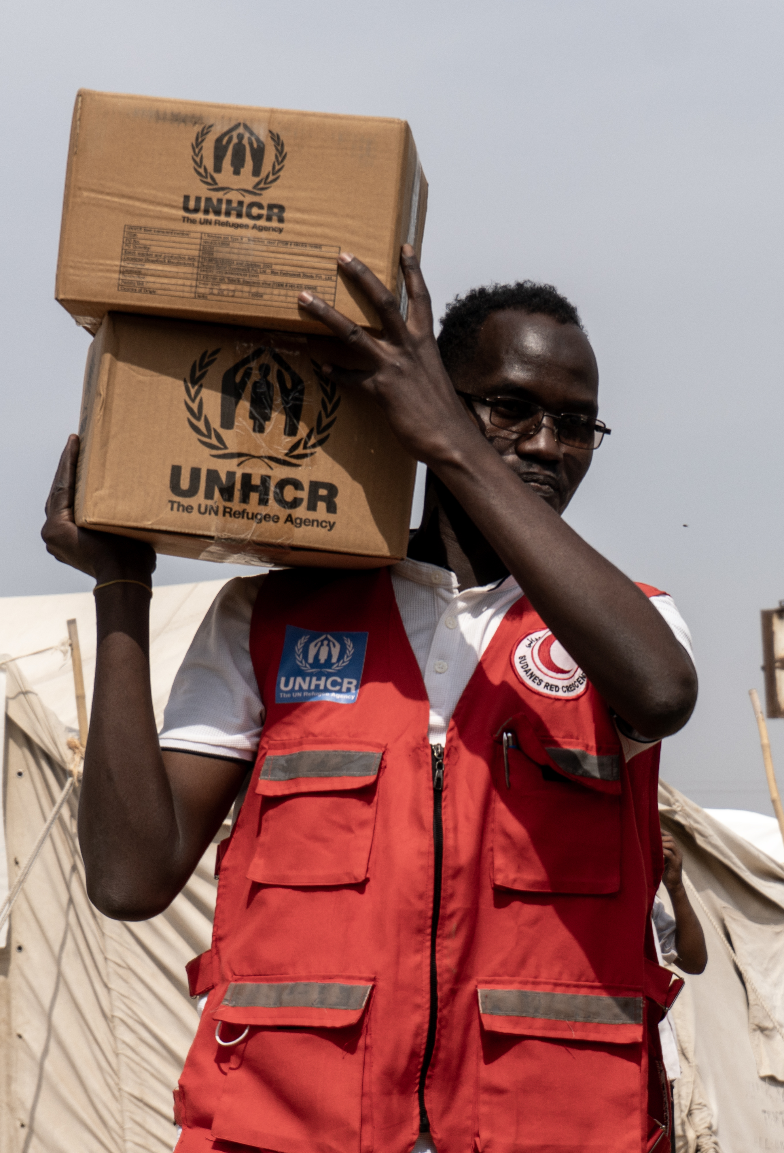 En mann i rød vest med logoene til UNHCR og Røde Halvmåne bærer to pappesker merket UNHCR på skulderen. Telt står i bakgrunnen.