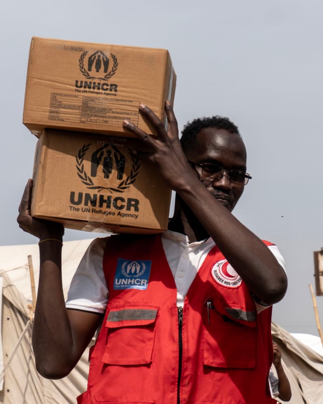 En mann i rød vest med logoene til UNHCR og Røde Halvmåne bærer to pappesker merket UNHCR på skulderen. Telt står i bakgrunnen.