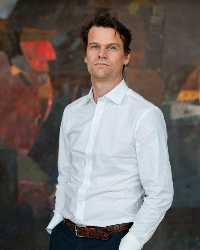 Portrett av en voksen mann i hvit skjorte og mørke bukser, stående foran en mørk, abstrakt bakgrunn. Han ser rett mot kameraet med den ene hånden i lommen.