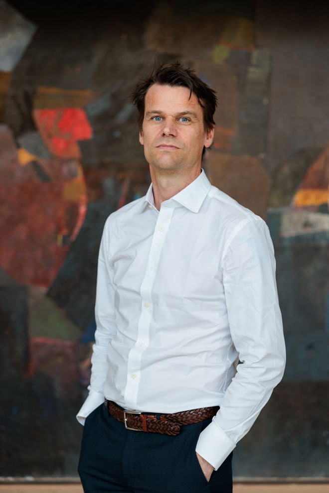 Portrett av en voksen mann i hvit skjorte og mørke bukser, stående foran en mørk, abstrakt bakgrunn. Han ser rett mot kameraet med den ene hånden i lommen.