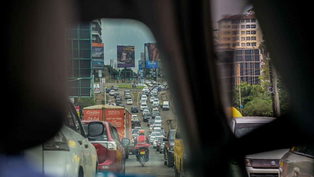 Kø av biler fyller en flerfelts vei i Nairobi, sett gjennom vinduene i et kjøretøy, mens en motorsykkel kjører mellom feltene. Bygninger, trafikklys og reklameskilt er synlige langs veien.